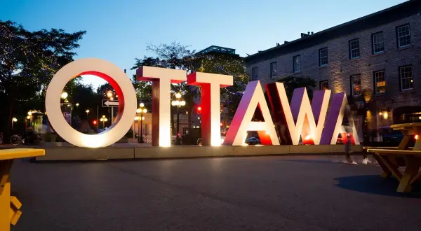 Napis OTTAWA, stolica Kanady, świeci w nocy. W tle budynki i drzewa ozdobione lampkami.