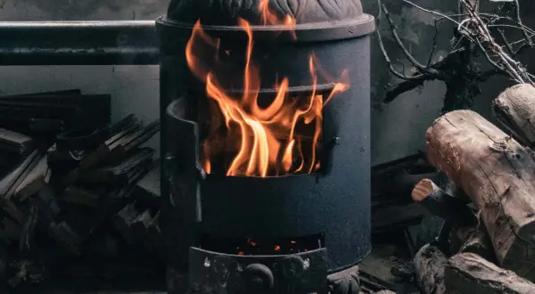 Ein Kaminofen brennt hell. Achte auf ausreichende Belüftung, um Sauerstoffmangel durch Kaminofen zu vermeiden. Holzscheite liegen daneben.