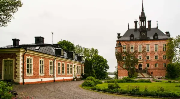 Vackert vik slott med en lång, röd tegelbyggnad och ett torn med klocka. Grönskande trädgård och en grusväg leder fram till byggnaderna.