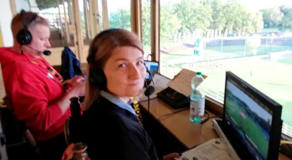 Komentator sportowy w słuchawkach przygotowuje się do transmisji. Obok monitor, mikrofon i butelka wody.