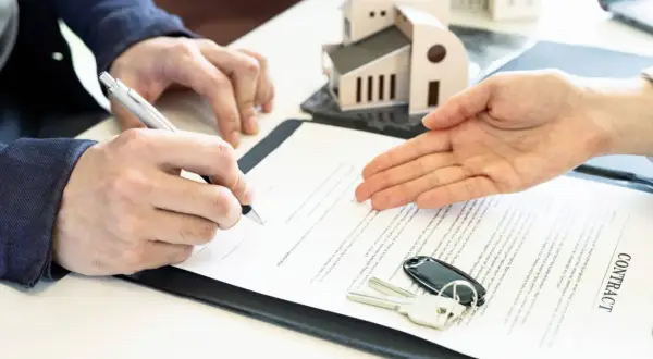 Signature d'un contrat immobilier. Un homme signe un document, des clés et un modèle de maison sont posés sur la table. Le courrier sru est prêt.