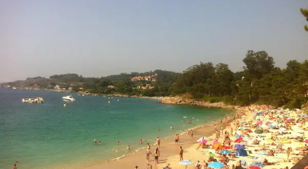 Gente disfrutando del sol y el mar en la playa areacova, con barcos en el agua y colinas verdes al fondo.