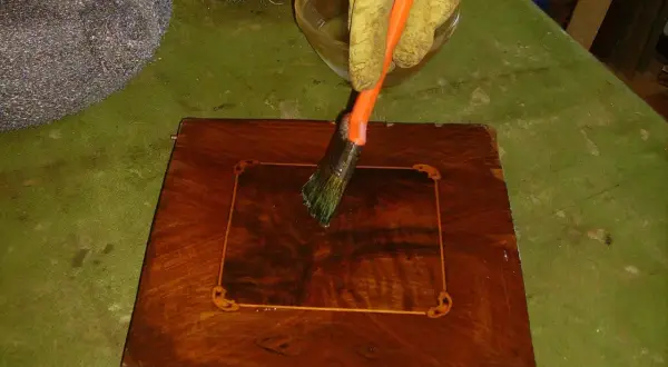 Mano guantata applica liquido con pennello per sverniciare mobile legno. Accanto, paglietta metallica e spazzola.