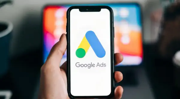 Dłoń trzyma smartfon z logo Google Ads. W tle widać ekran laptopa. Dziś budżet w reklamie Google Ads ustalamy.