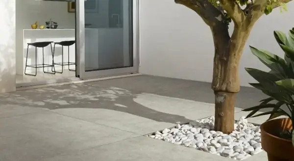 Un moderno patio con diversi tipi di pavimentazione esterna: lastre in gres grigio e un'area con ciottoli bianchi attorno a un albero.