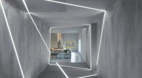 Nowoczesny korytarz z betonowymi ścianami i sufitem, ozdobiony geometrycznymi liniami światła LED. W tle widać salon z kominkiem i meblami. To inspiracja, jak zamontować oświetlenie LED w witrynie.