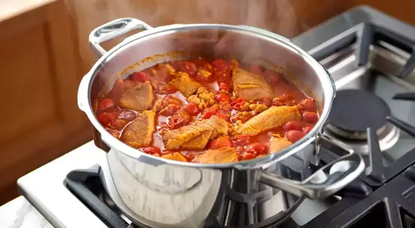 Un délicieux saute de porc mijote dans une cocotte en acier inoxydable sur une cuisinière à gaz, dégageant une vapeur appétissante.