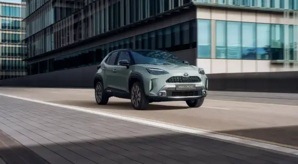 En grön Toyota Yaris Cross hybrid kör på en stadsgata. Bilen är i fokus framför moderna byggnader.