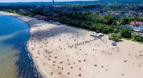 Słoneczny dzień, plaża pełna ludzi, idealne miejsce, gdzie pociągiem nad morze. W tle widać miasto i zieleń.