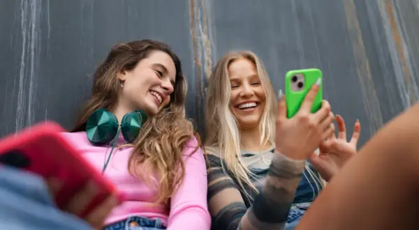 Dwie uśmiechnięte dziewczyny rozmawiają przez **Facetime** na telefonach, jedna z nich trzyma zielony smartfon.