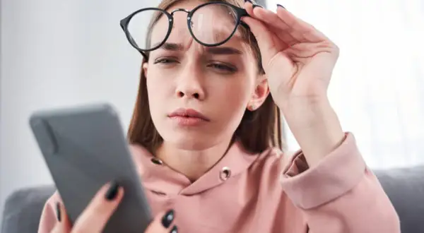 Zmarszczona kobieta w okularach trzyma telefon, szukając domowych sposobów na mroczki przed oczami.