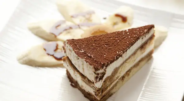 Tranche de tiramisu banane, saupoudrée de cacao, accompagnée de rondelles de banane nappées de sauce. Un délice !