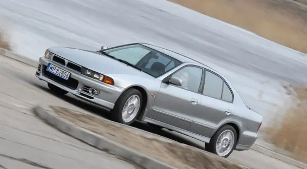 Srebrny Mitsubishi Galant z kierowcą za kierownicą, pokonujący zakręt na drodze.