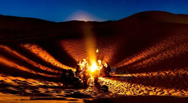 Un groupe se réchauffe autour d'un feu de camp dans le désert du Sahara la nuit. La température chute, mais la chaleur du feu et la beauté du ciel étoilé créent une atmosphère magique.