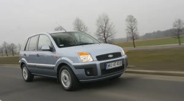 Jasnoszary Ford Fusion 1.4 benzyna jedzie po drodze. W tle drzewa i pola.