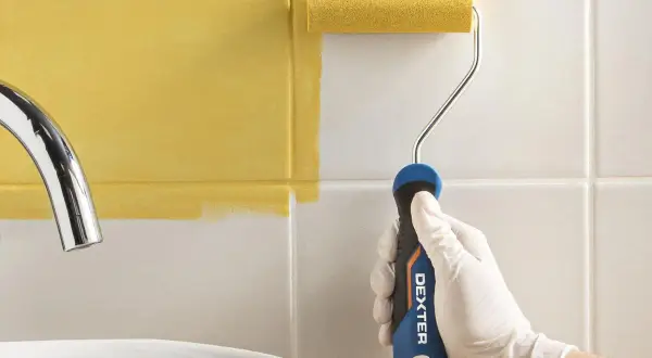 Mano con guante blanco pintando azulejos de baño de blanco a amarillo con rodillo.