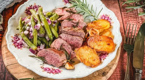 Saftiger Lammbraten nach Omas Rezept, serviert mit grünen Bohnen, roten Zwiebeln und knusprigen Kartoffelscheiben auf einem Teller mit Blumenmuster.
