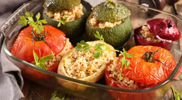 Un plat de petits farcis gastronomiques : tomates, courgettes rondes et oignons rouges farcis de grains et herbes, dans un plat en verre.