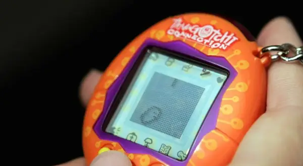 Pomarańczowe Tamagotchi Connection w dłoni, z widocznym ekranem i przyciskami. Pamiętasz, co to tamagotchi?