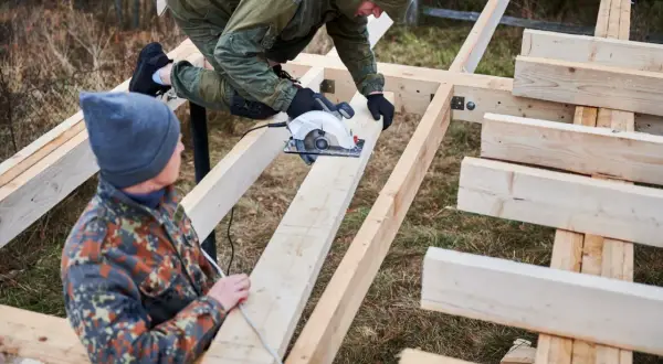 Dwóch mężczyzn buduje konstrukcję z drewna. Jeden z nich używa piły tarczowej do cięcia belek, które są częścią projektu.