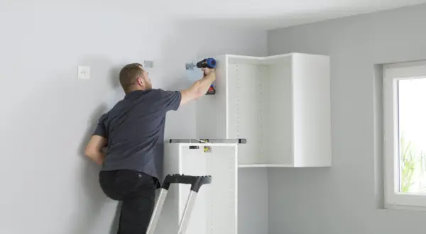 Hombre en escalera usa taladro para fijar soportes en pared, parte de como colocar kit para colgar armarios.