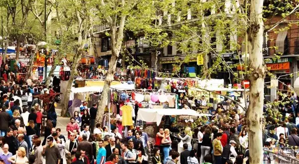 Gente comprando en mercadillos de la sierra de Madrid, con puestos de ropa y accesorios bajo árboles frondosos.