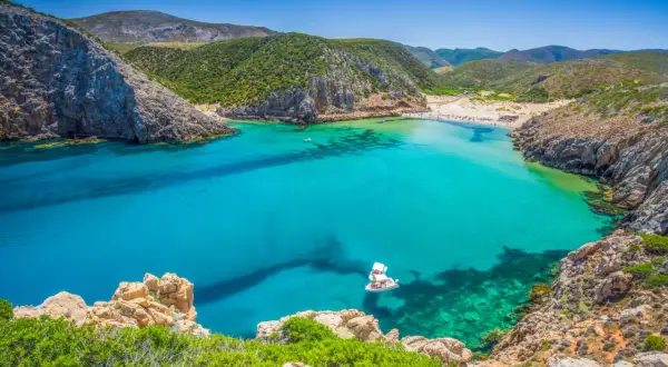 Traumhafte Cala Domestica mit türkisfarbenem Wasser, Felsen und grünen Hügeln. Ein Boot ankert in der Bucht.