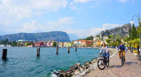 Coppia in bici sul lungolago di Riva del Garda, tra le attrazioni del posto, con case colorate e montagne sullo sfondo.