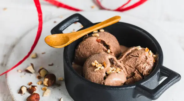Schokoladiges Nutella-Eis mit 3 Zutaten, garniert mit gehackten Haselnüssen in einer schwarzen Schale, daneben eine Holz-Löffel und rote Bänder.