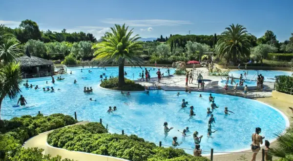 En stor pool med många badande människor, palmer och en vattenrutschkana. Perfekt för camping i Italien.