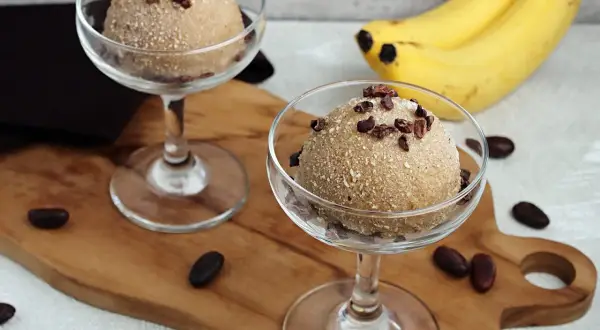 Zwei Gläser mit cremigem Apfel Zimt Eis, garniert mit Kakaonibs. Bananen im Hintergrund.