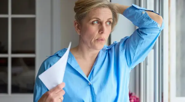 Kobieta w niebieskiej koszuli wachluje się kartką, czując uderzenia gorąca. Czy to już moment, kiedy ustępują objawy menopauzy?