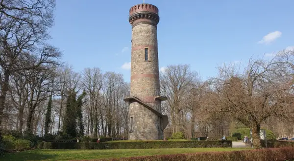 Der Toelleturm in Wuppertal, ein steinerner Aussichtsturm, ragt vor kahlen Bäumen und blauem Himmel auf. Eine Wendeltreppe führt zur Aussichtsplattform.