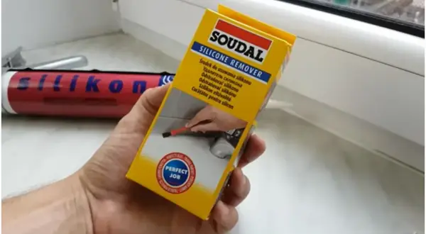 Ręka trzymająca opakowanie środka do usuwania silikonu Soudal. W tle widać tubkę z silikonem.