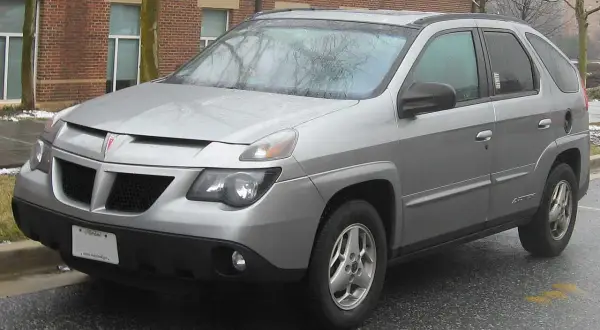 Srebrny Pontiac Aztek zaparkowany na mokrym asfalcie, z ceglanym budynkiem w tle.