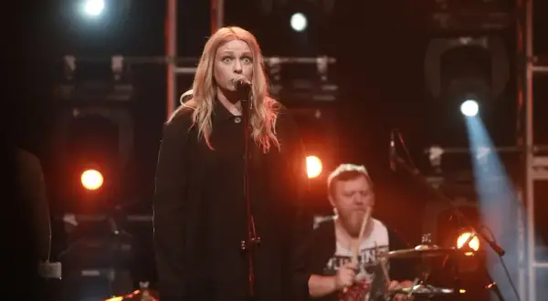 Wokalistka z blond włosami śpiewa do mikrofonu, a za nią perkusista gra na bębnach. To jak hey teksański tekst na scenie.