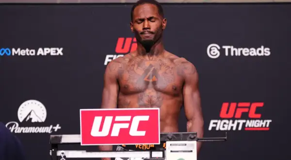 A fighter with tattoos stands on a scale, eyes closed, preparing for weigh-ins. The scale's weight brackets are visible.