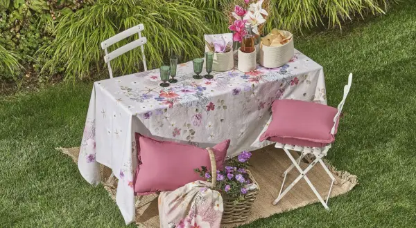 Un tavolo da giardino apparecchiato con una tovaglia floreale, bicchieri verdi, cesti e cuscini rosa. Un'idea per come rivestire un tavolo con stoffa per un picnic.