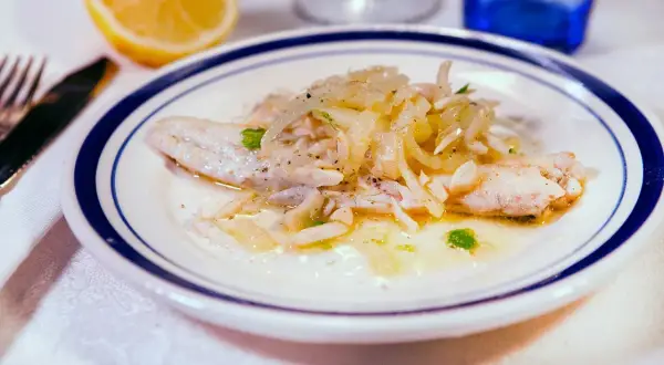Delicato branzino in padella con cipolle e prezzemolo, servito su un piatto bianco con bordo blu. Accanto, uno spicchio di limone.