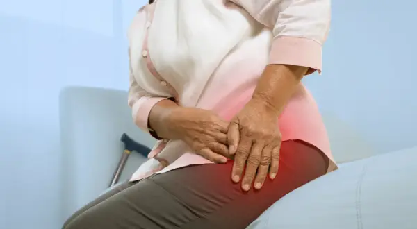 Une femme âgée se tient la hanche, souffrant de mal de hanche et sciatique. Une canne est visible derrière elle.