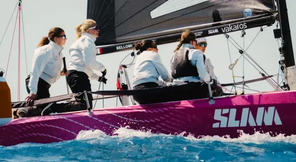 Equipaggio femminile su barca a vela rosa, con la vela nera e il logo SLAM. Le veliste si preparano per la regata RS 21.