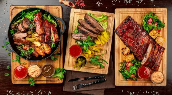 Trzy deski z grillowanym mięsem: żeberka, steki i skrzydełka kurczaka, podane z frytkami, kukurydzą i sosami.