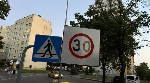 Znak D43 (przejście dla pieszych) i znak ograniczenia prędkości do 30 km/h na tle bloków mieszkalnych i drzew.
