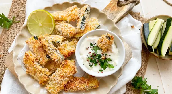 Knusprige Zucchini Pommes, paniert und goldbraun gebacken, serviert mit Dip und Limette.