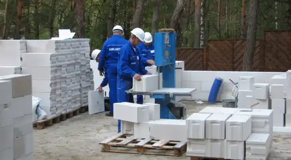 Budowa domu z bloczków betonowych. Pracownicy w niebieskich kombinezonach i białych kaskach układają materiał. Widać piłę do cięcia bloczków.