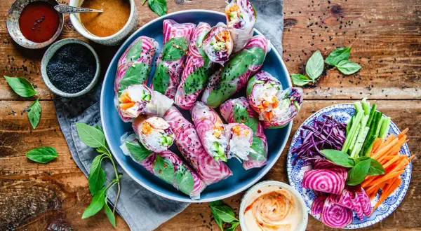 Färgglada vietnamesiska vårrullar med grönsaker och risnudlar, serverade med dippsåser och färska örter på en träbakgrund.