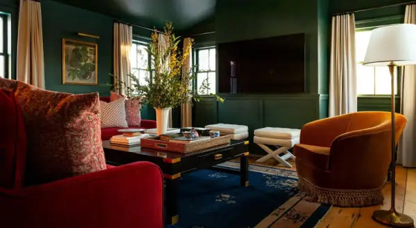 A rich, jewel-toned living room with plush red sofas, an orange velvet armchair, and a dark coffee table, showcasing timeless interior design.