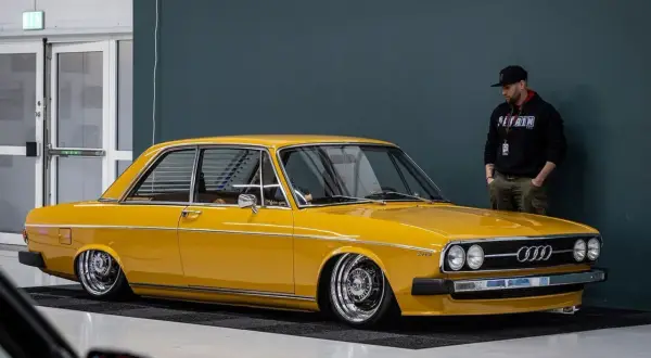 Gul Audi 100 C1, en klassiker med kromade fälgar, står parkerad bredvid en man i keps.