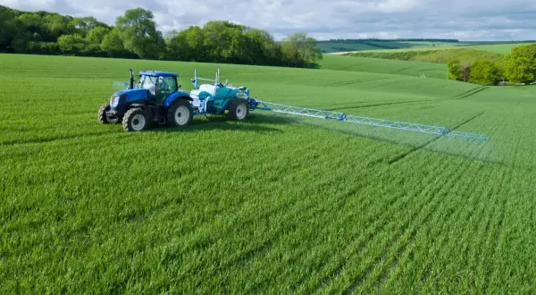 Niebieski traktor z opryskiwaczem na zielonym polu, stosuje środek Chwastox Turbo 340 SL. Dawkowanie jest kluczowe dla skuteczności.