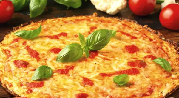 Pyszna pizza z kalafiora, z ciągnącym się serem, pomidorami i świeżą bazylią. Obok leżą pomidory i różyczki kalafiora.